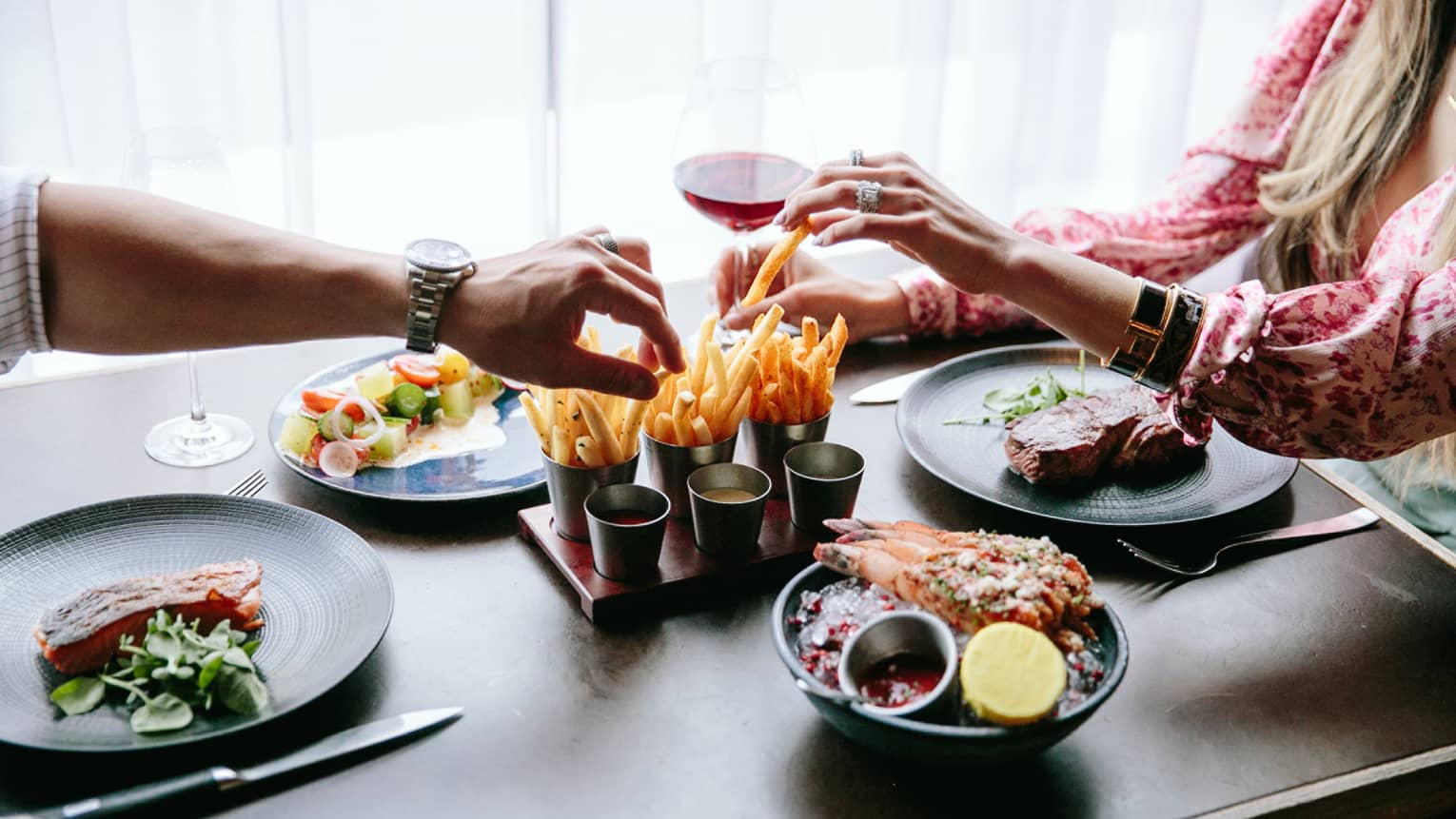 Two people eating an assortment of fries, steak, and salad.