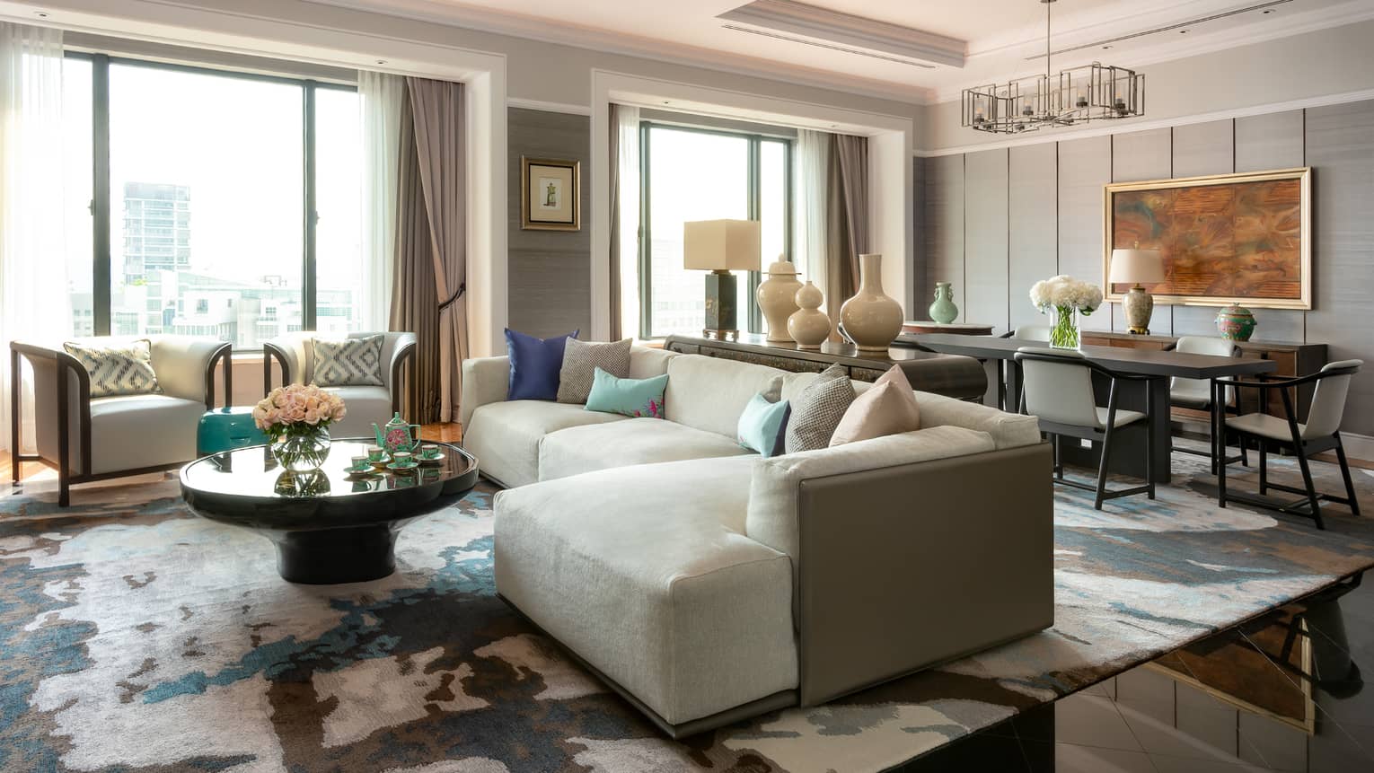 A spacious, luxurious living room with large windows, light grey sectional sofa, round coffee table and modern décor, including vases and artwork on the walls.