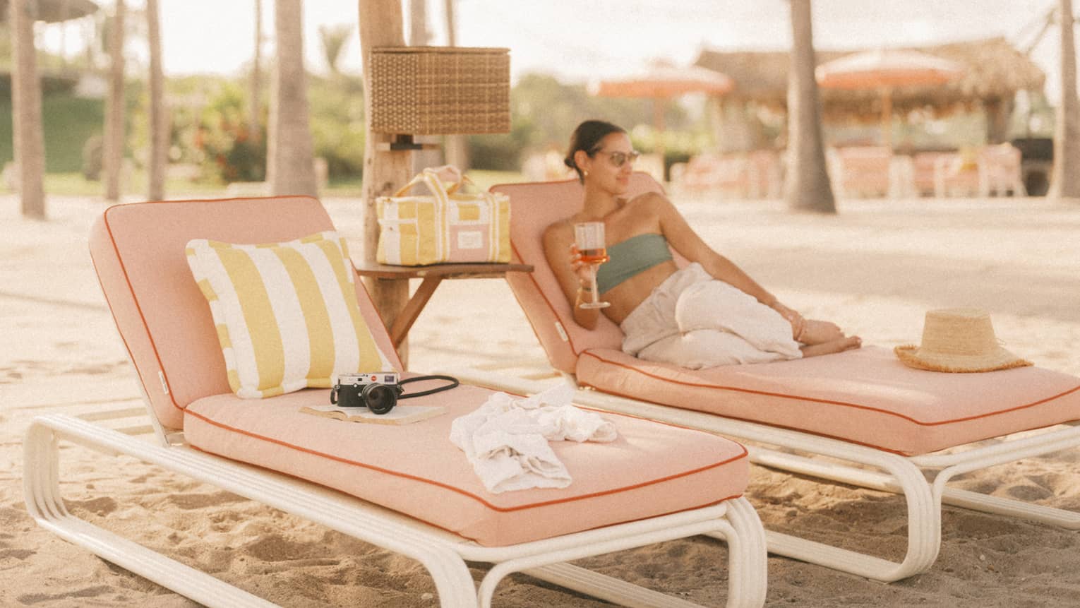 Two lounge chairs on a beach, side by side under a thatched umbrella for shade. A person reclines, drink in hand, on one of the chairs.