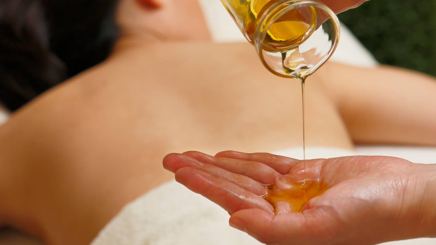 Spa staff pouring massage oil from glass jar in hand over woman lying on table