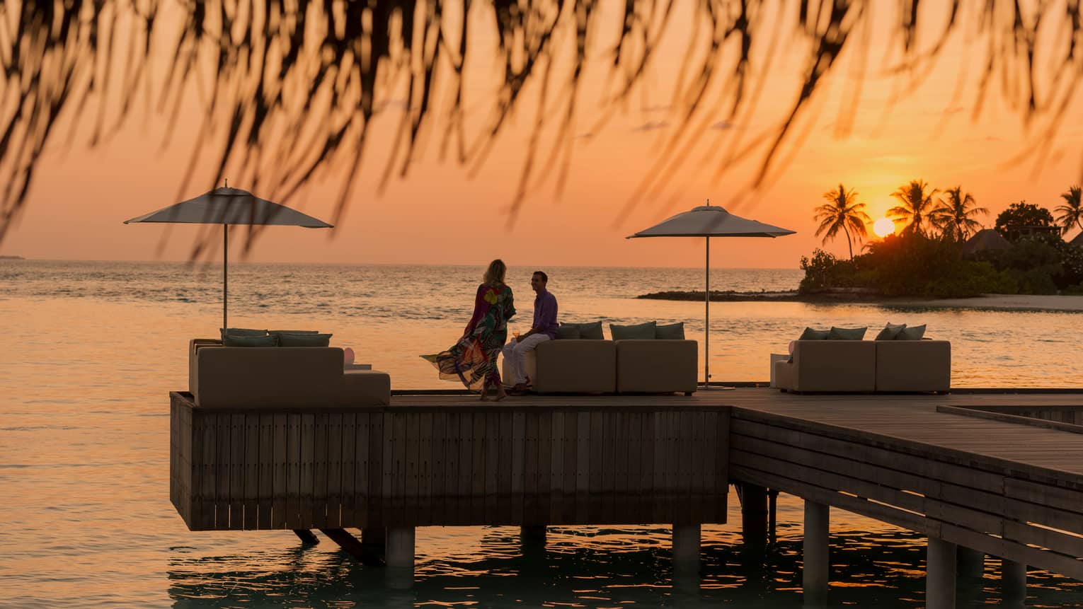 Private island dining at sunset. Woman with long dress blowing in the wind, man sits on the edge of white sofa