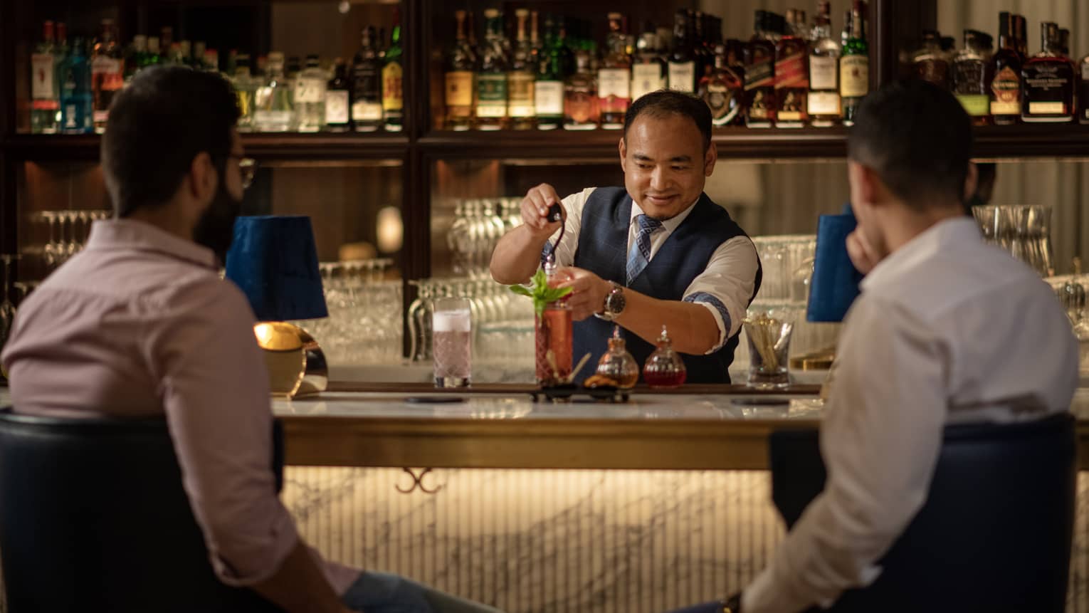 A bartender making a cocktail while two men converse at a bar.