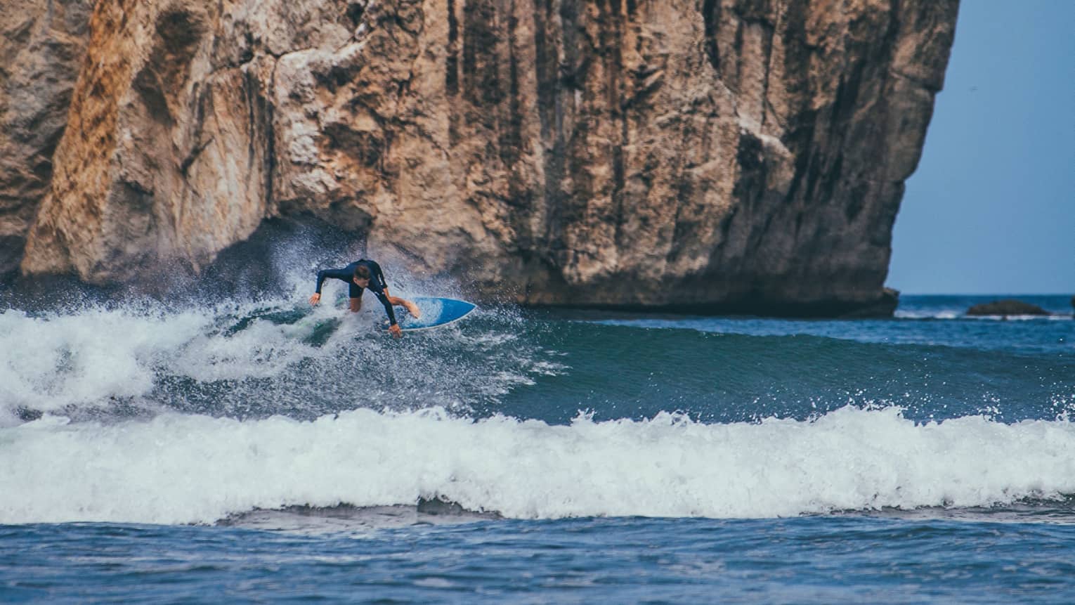 A surfer rides the crest of a wave, body angled forward, amid frothy sea foam; a giant ochre cliff looms in the background.