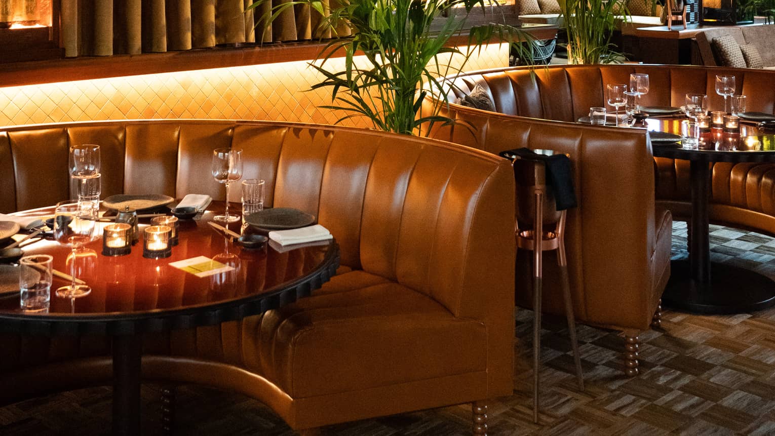 Several brown leather loveseats in front of circular tables in a dimly lit restaurant.