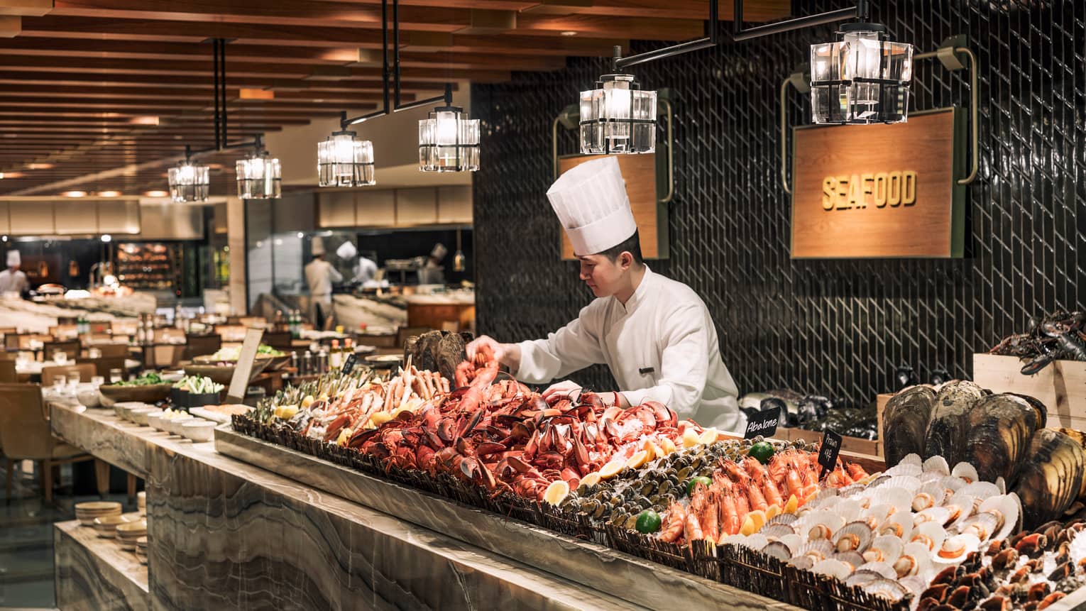 Chef wearing tall white hat arranges fresh lobster, seafood on ice at buffet The Market Kitchen buffet station