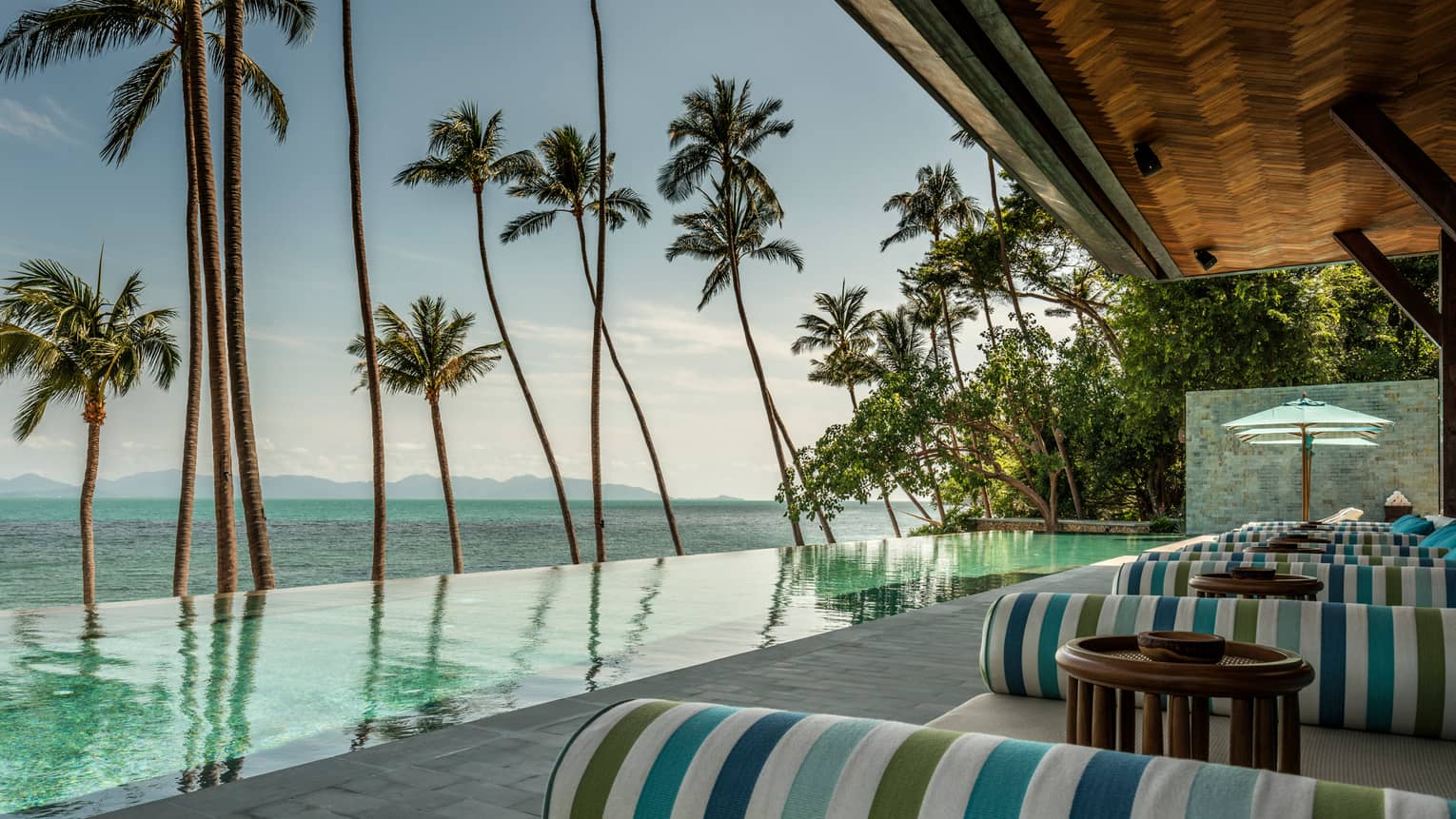 Green-and-blue striped banquettes on patio by infinity swimming pool, ocean