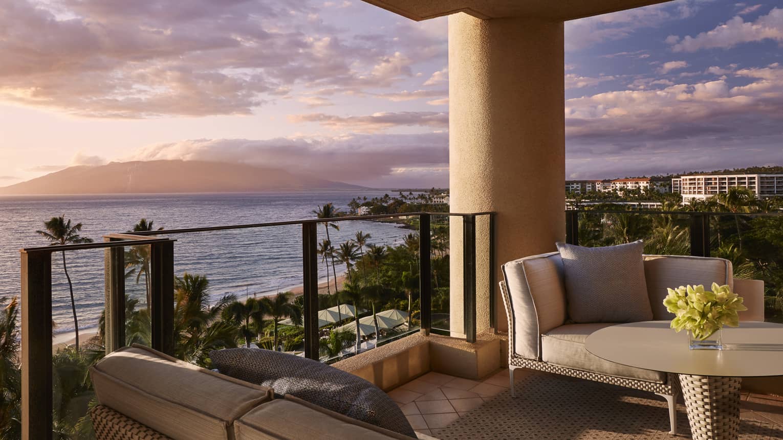 Balcony with cushioned seating and table overlooking beach palm trees and ocean at sunset