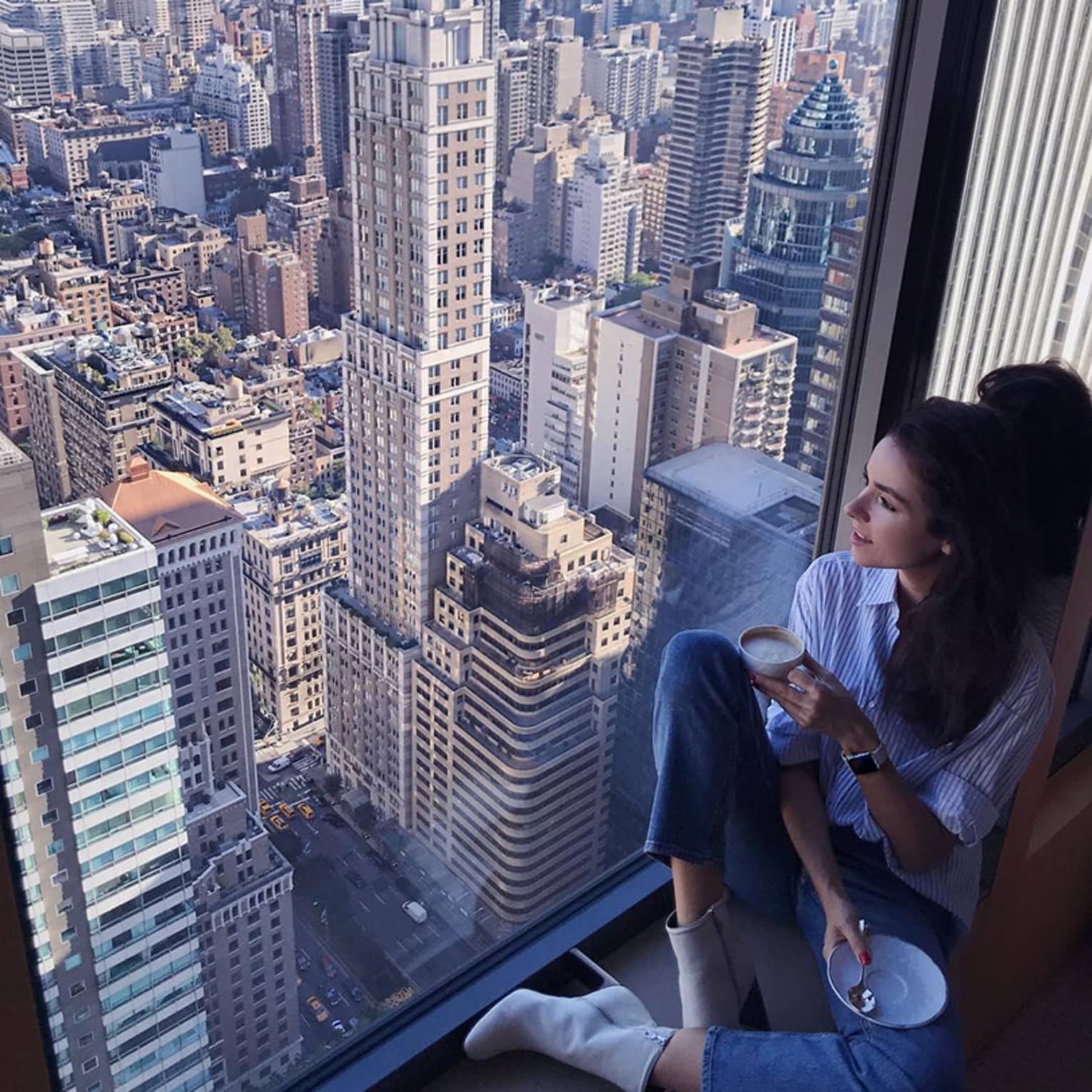 Woman with coffee cup sits in large window overlooking New York City rooftops
