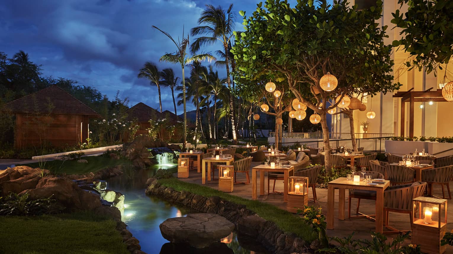 Noe patio dining tables, chairs, lanterns by stream at night