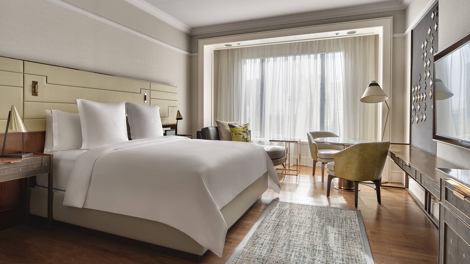 Light-filled hotel room with white king bed, wood floors and rug, two arm chairs, sheer curtains