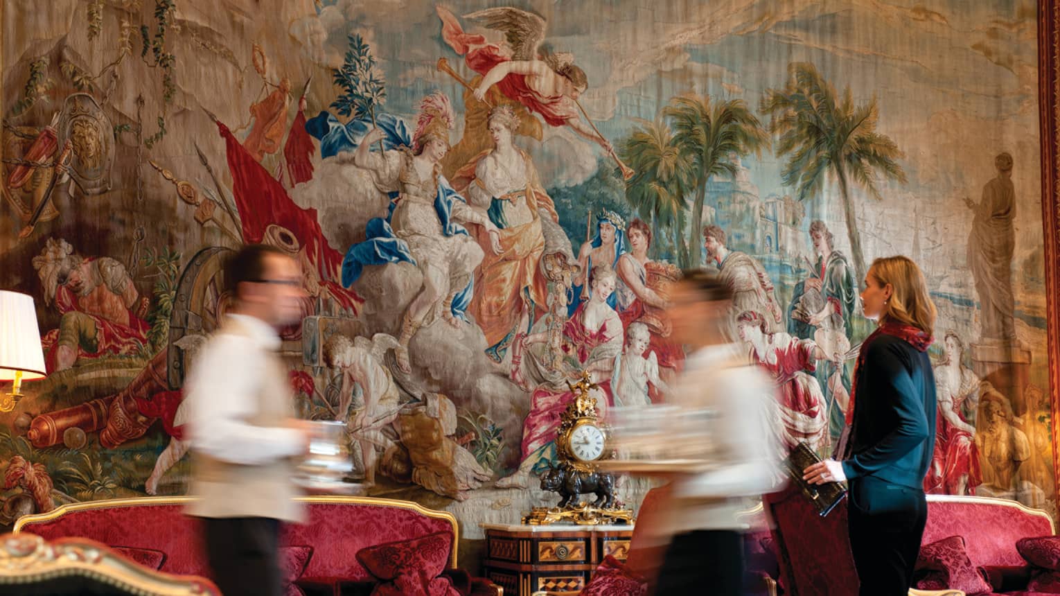 Servers walk by antique red velvet sofas, large wall textile