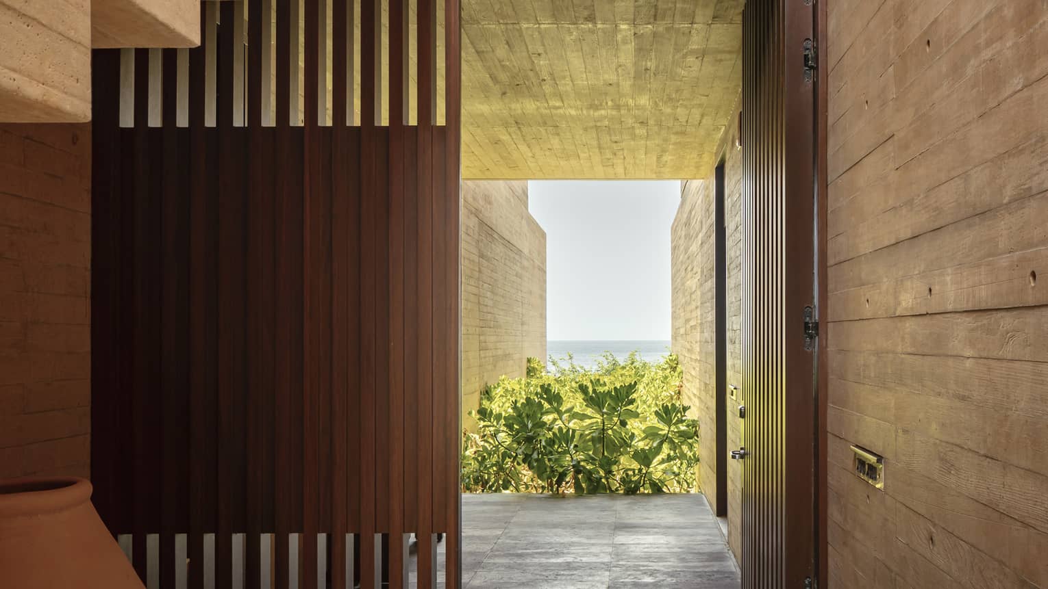 A modern, minimalist hallway with wooden walls and vertical slats. A staircase with metal railings leads to an upper level, while the lower passage opens to a garden with ocean views.