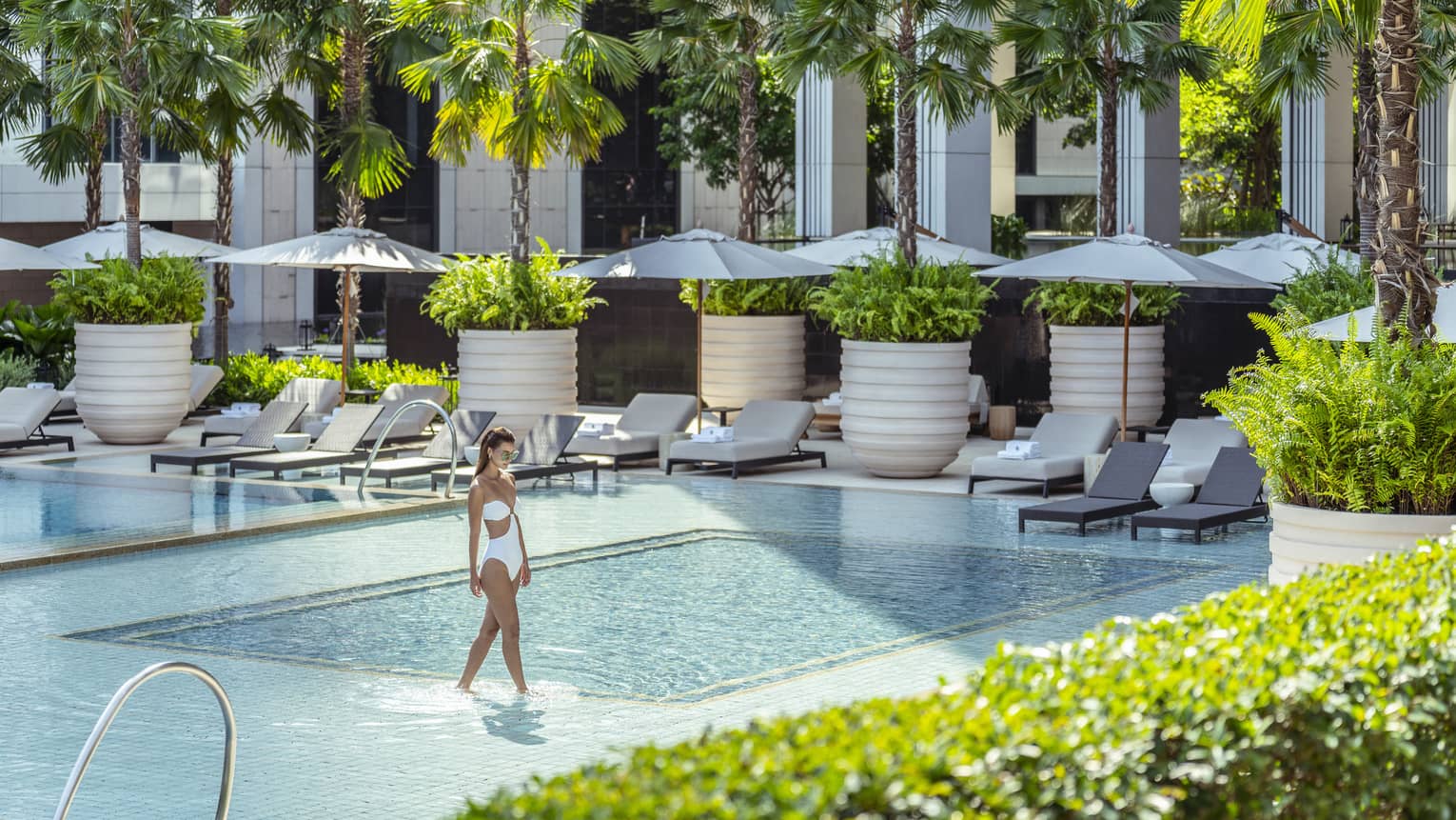 Woman in white bathing suit wades in outdoor pool surrounded by lounge chairs with umbrellas and palm trees