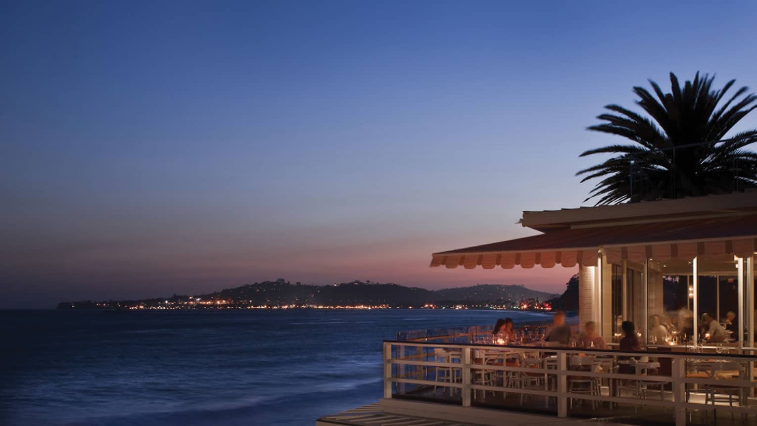 Diners on Tydes restaurant patio, palm tree silhouette, ocean at sunset