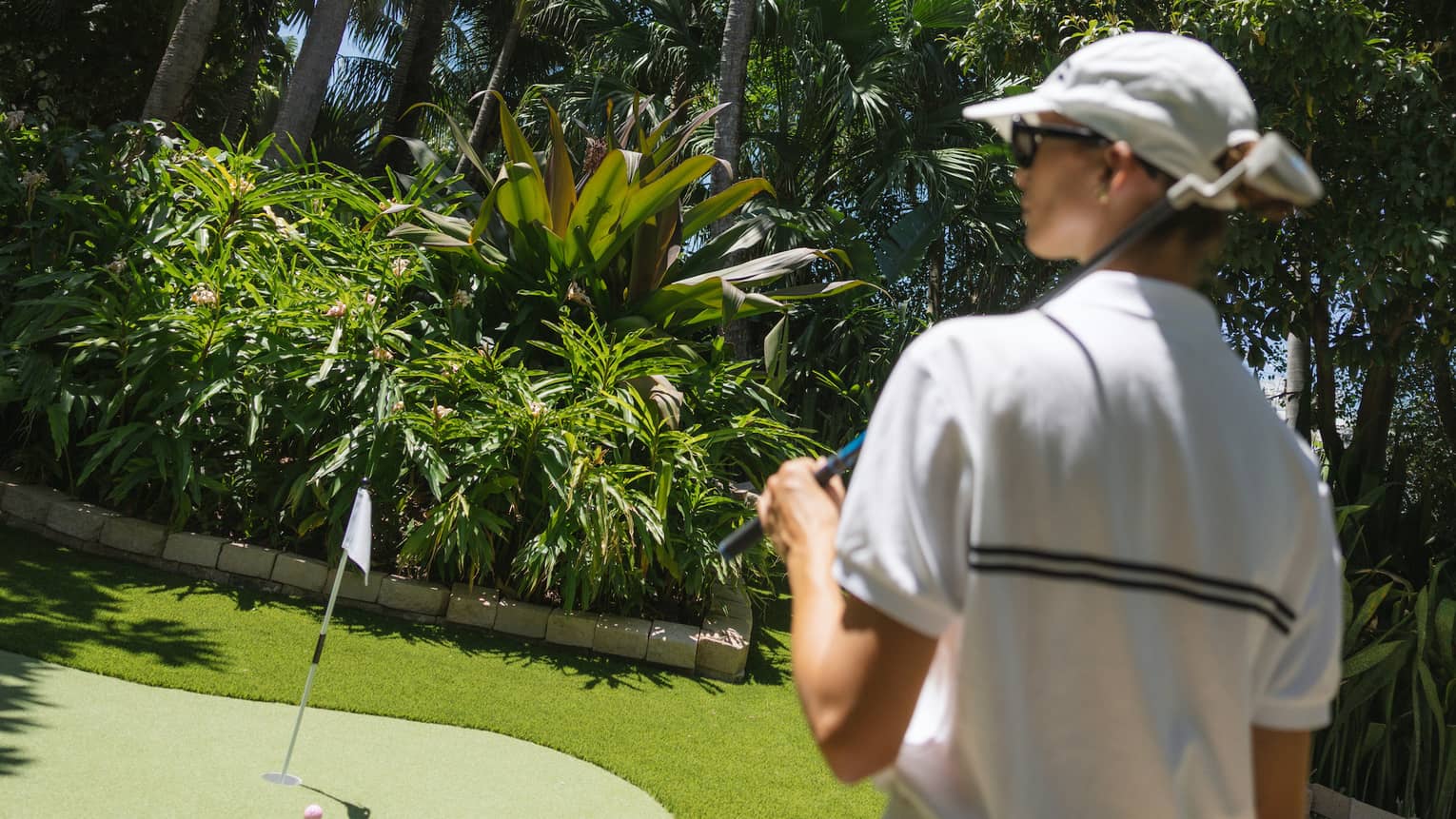Golfer wearing white sweater vest, white skirt and white hat walks toward a putting green surrounded by palm trees