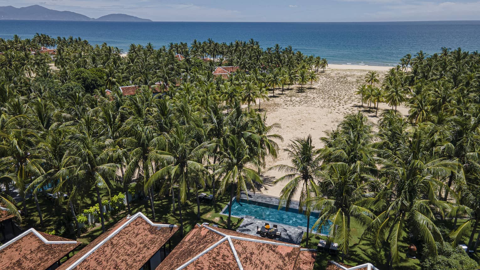 Aerial view of a beachfront Resort surrounded by lush palm trees, with a sandy beach, turquoise ocean and distant islands under a clear sky.