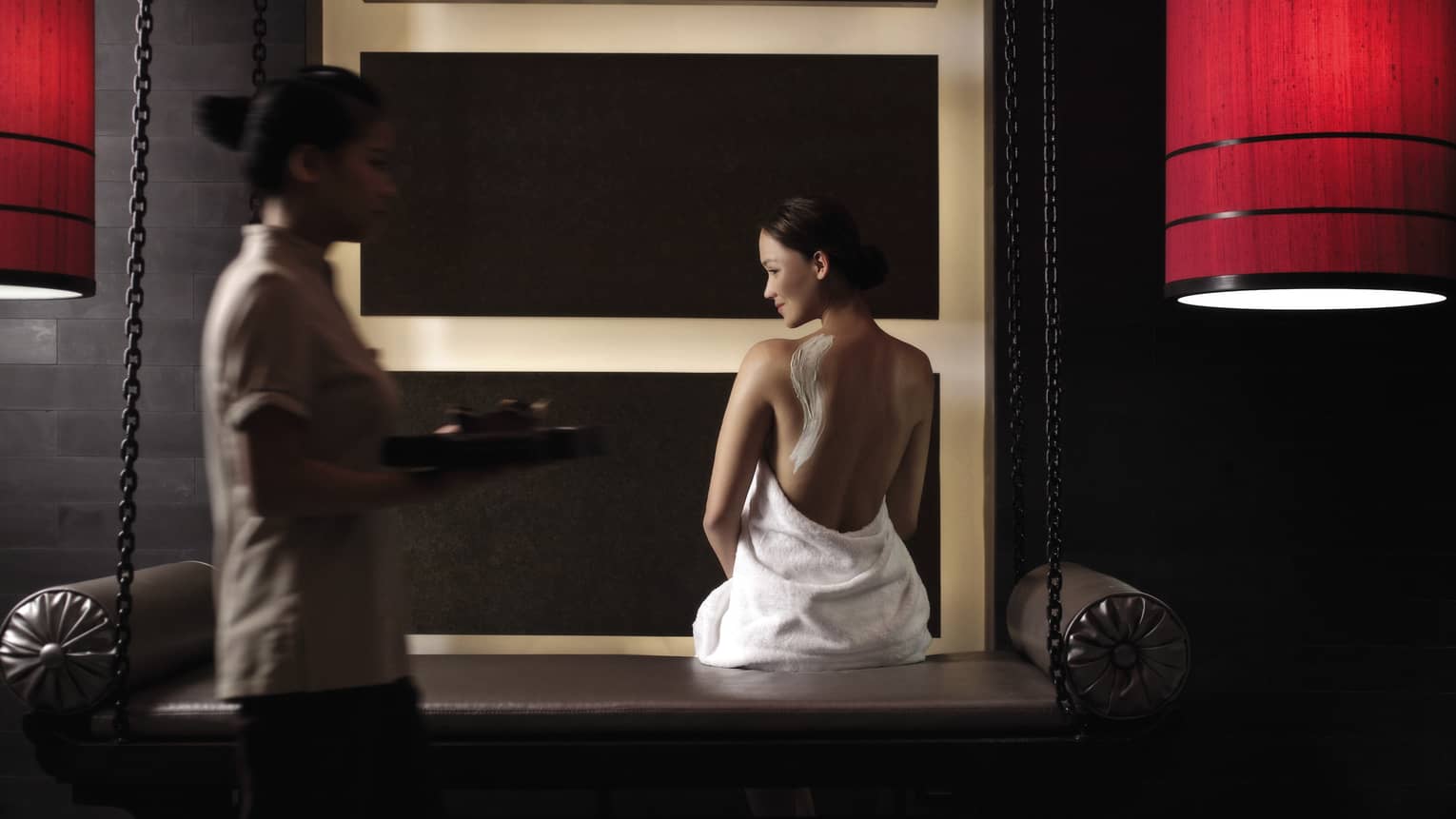 Woman in white towel with cream on back sits on bench in dark spa room