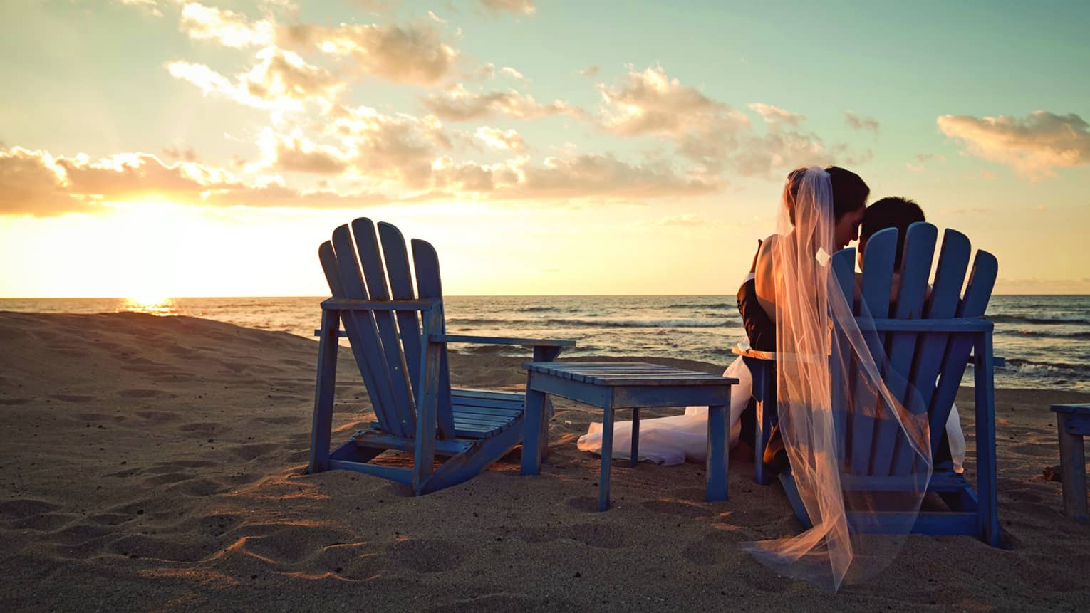 Back of bride and groom sharing chair on sand beach at sunset