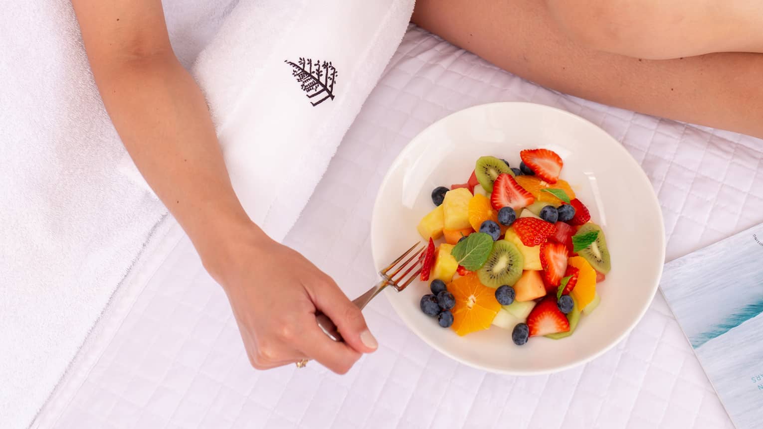 A woman in a blue swimsuit eats a bowl of colourful fruit while lounging in a cabana