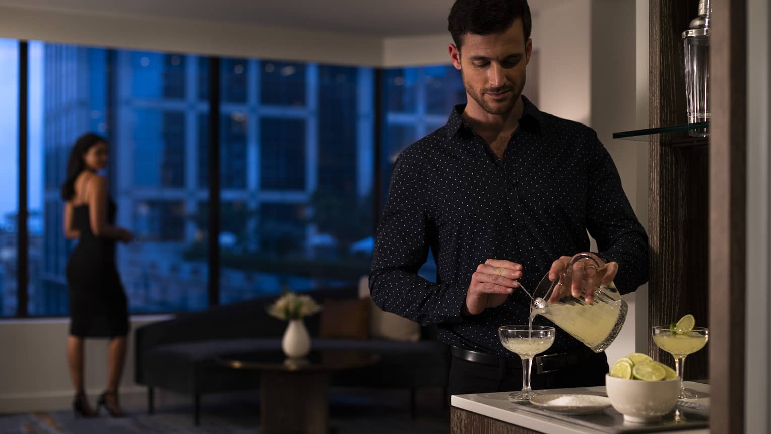 A man making a drink while a woman looks out a window.