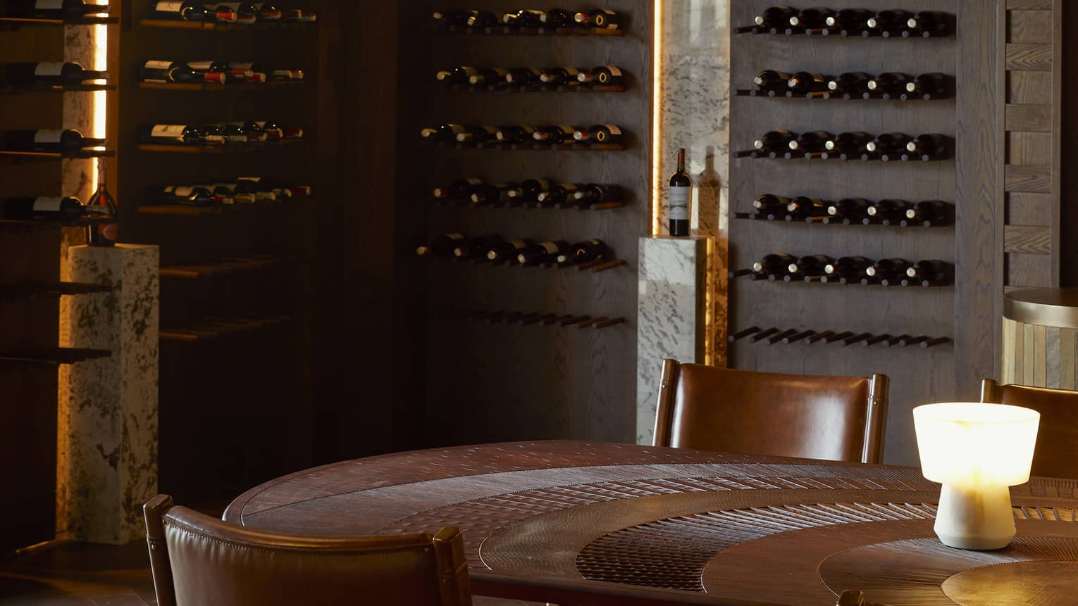 Dimly lit wine cellar with bottles lining dark wooden walls and round table in centre