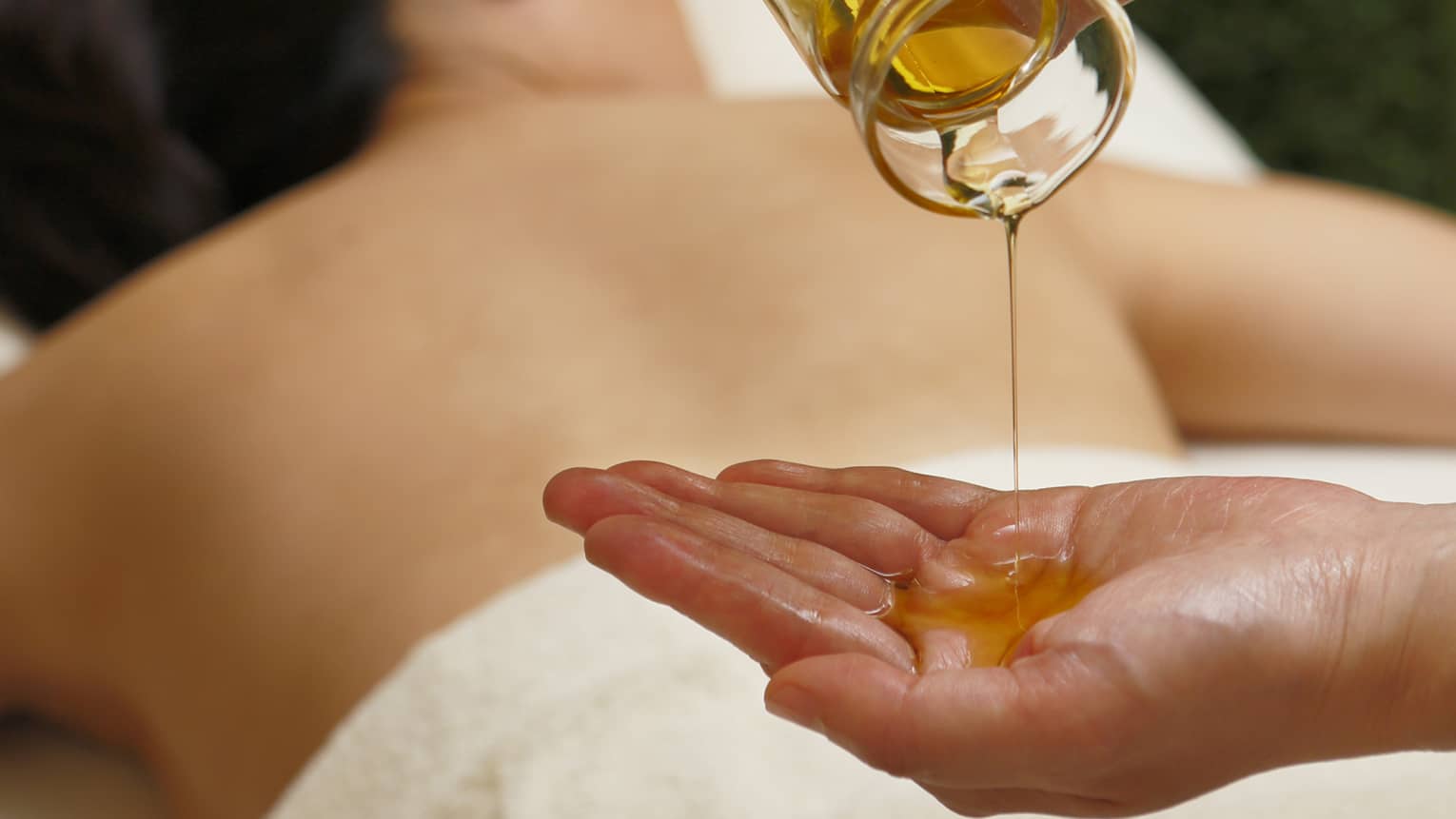 Spa staff pouring massage oil from glass jar in hand over woman lying on table