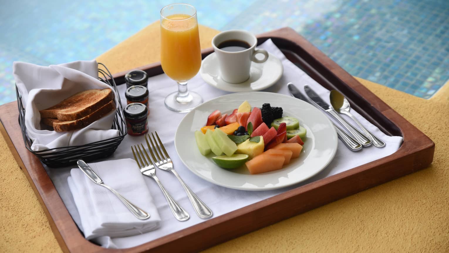 Fresh fruit slices on plate, coffee, orange juice, toast on wood tray by pool