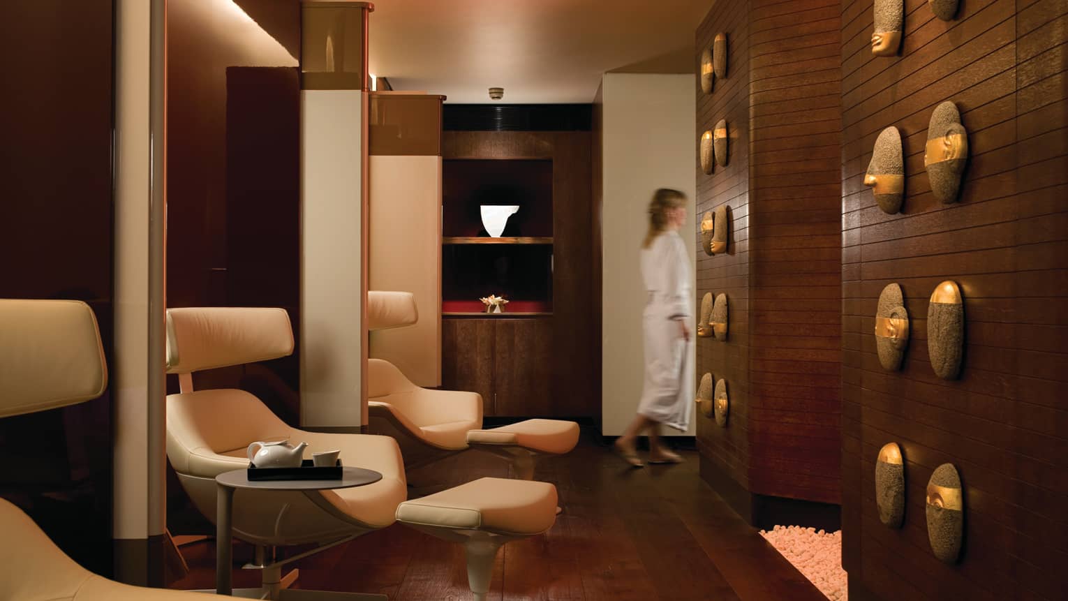 Woman wearing white bathrobe walks past modern wood panel wall, white leather chairs, tea trays