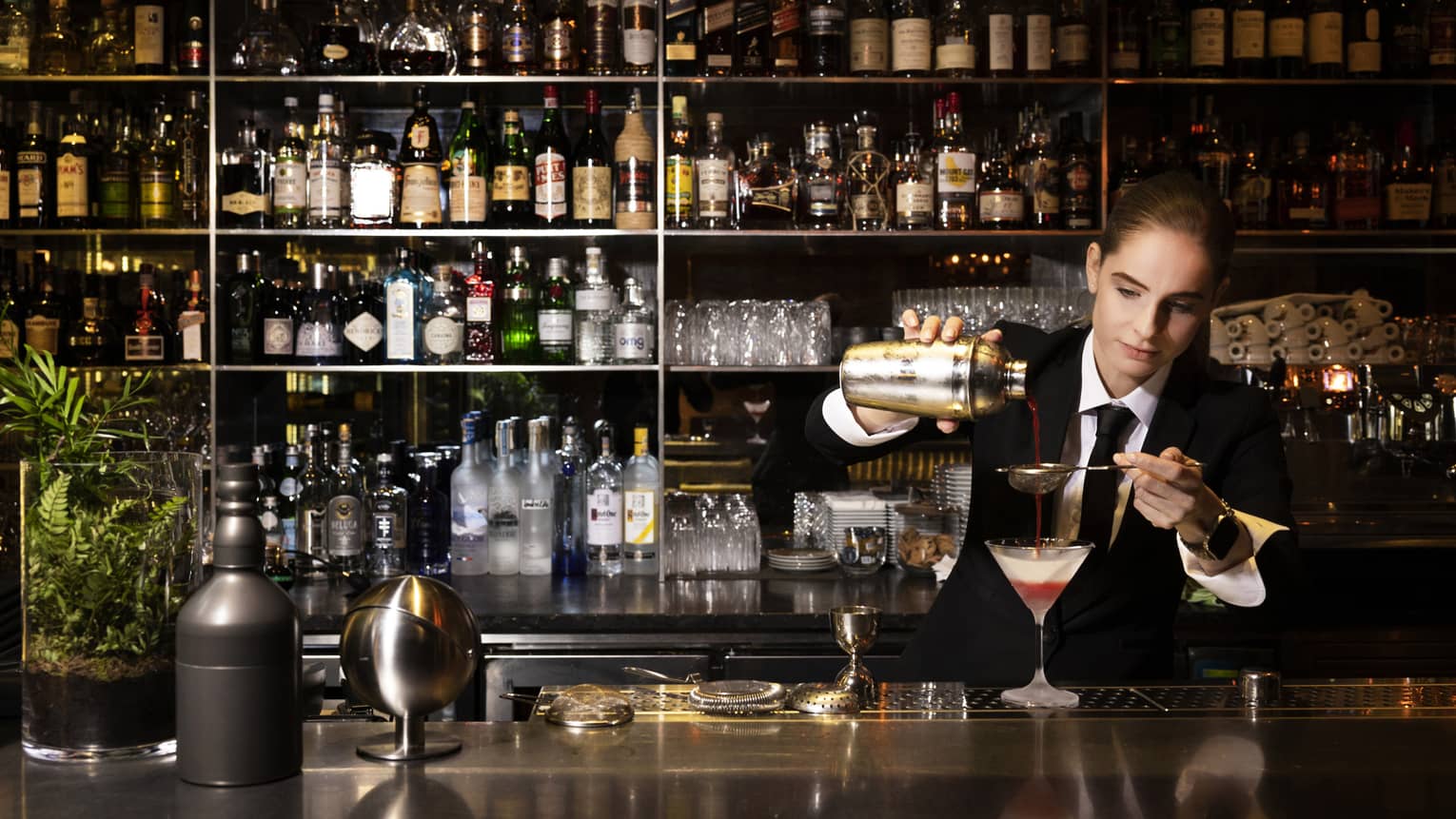 Bartender Silvie wearing a dark suit strains a red cocktail into a Martini glass on bar with stocked shelves behind