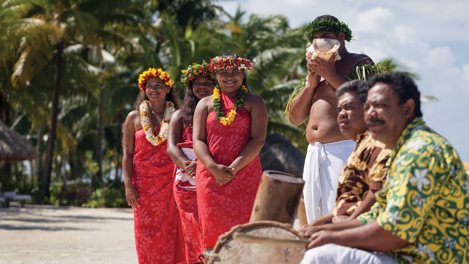 Group of people in traditional attire with leis and a conch shell, standing on a beach