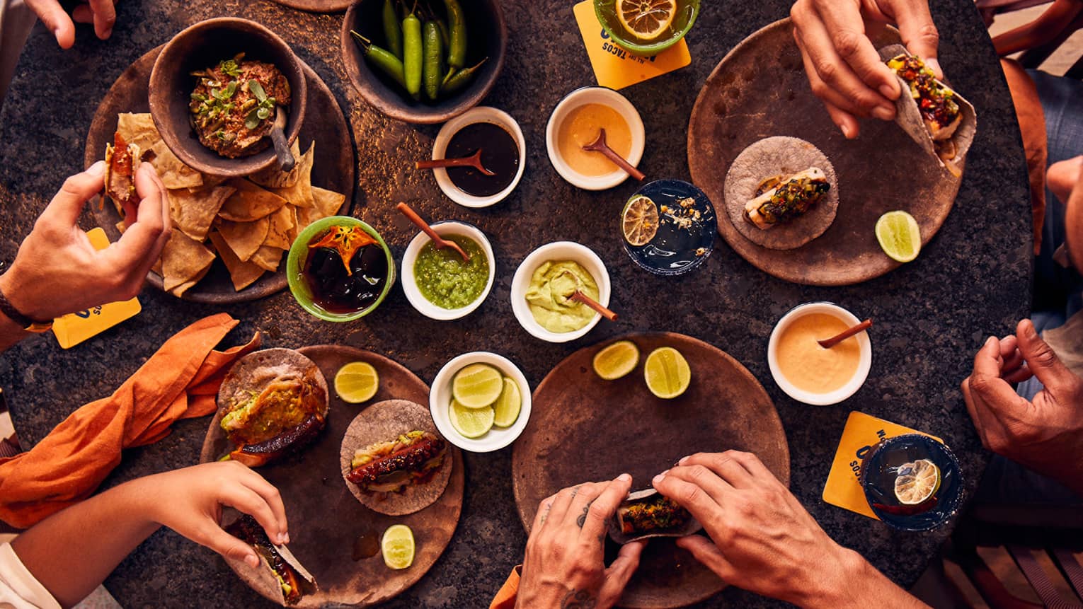 An overhead view of five pairs of hands holding tacos next to bowls of chips, salsa, peppers, limes and sauces.