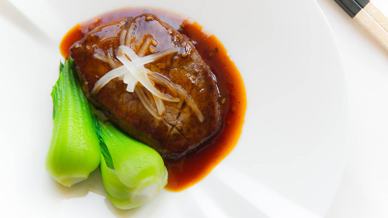 Beef topped with sauce and garnish with side of bok choy served on white plate