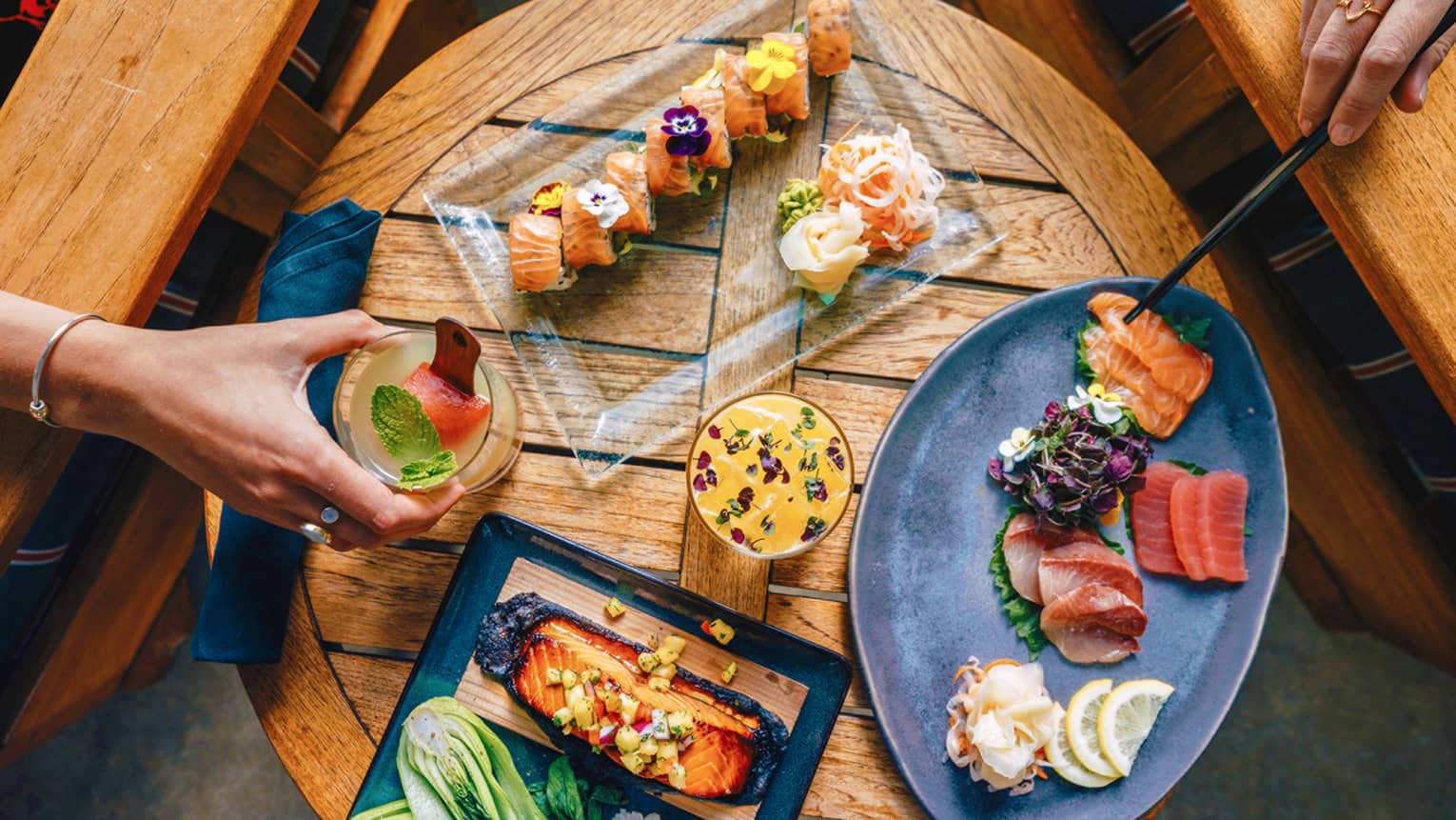 An aerial view of guests enjoying sushi and cocktails on a small, round wooden table 