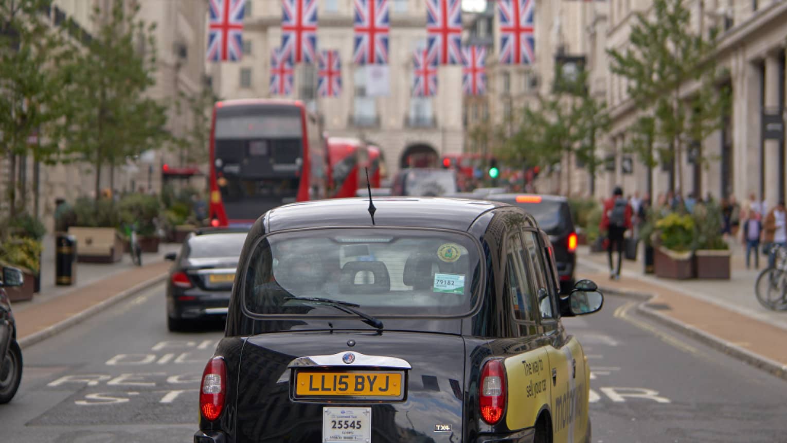 Rear view of London-style black cab, double-decker buses and other cars on road strung across with rows of Union Jack flags.