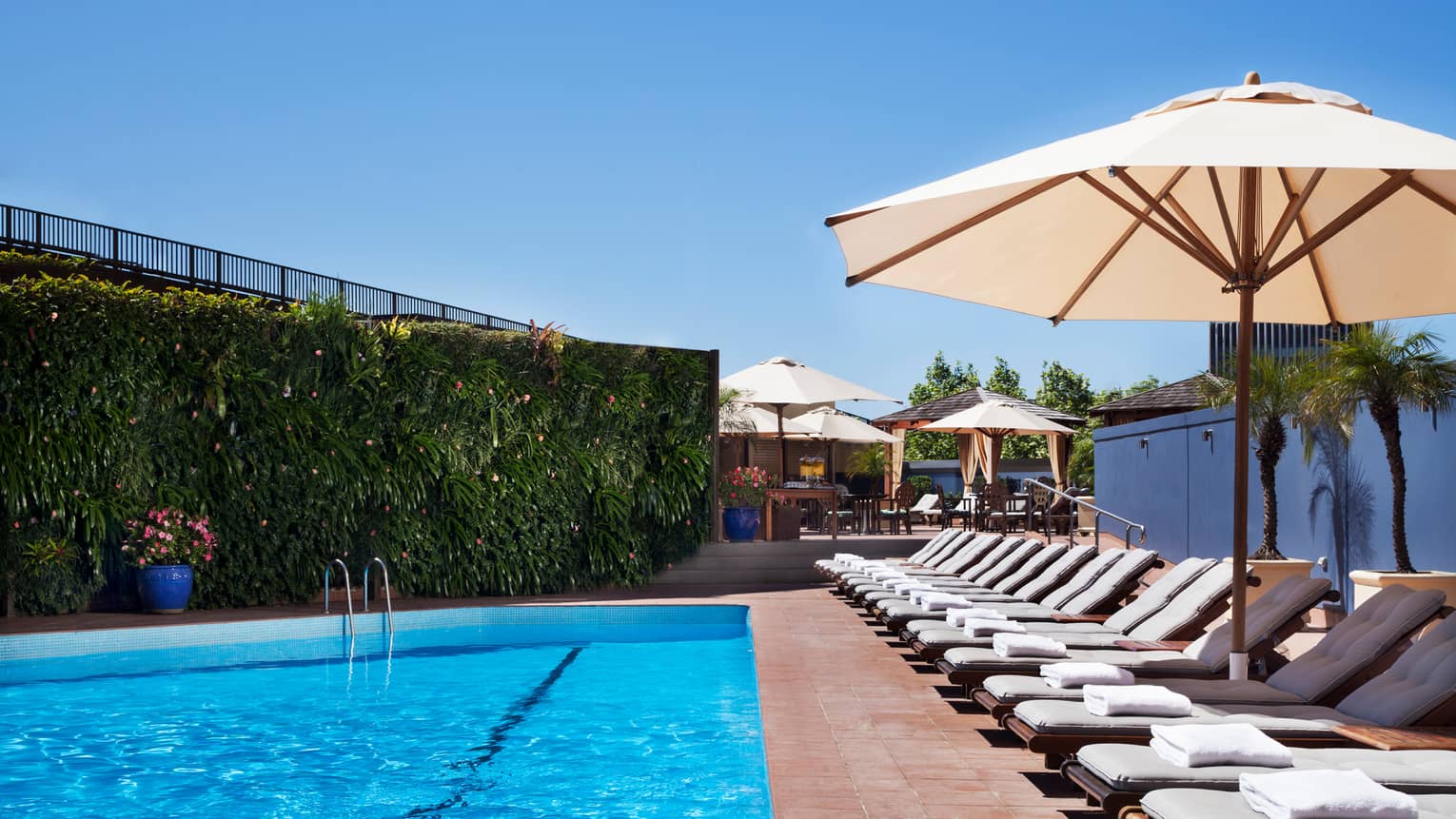 White lounge chairs lined up on deck under patio umbrella by blue outdoor swimming pool
