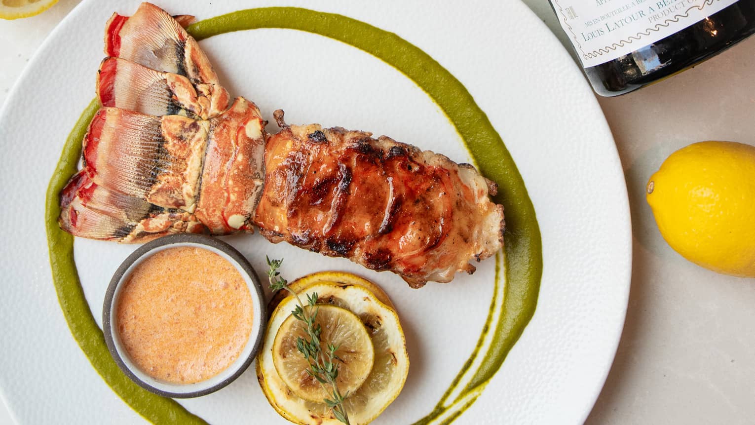 Plate of spiny lobster tail with a ramekin of butter and slices of torched lemon with a glass of wine, bottle of wine and lemon halves surrounding the plate