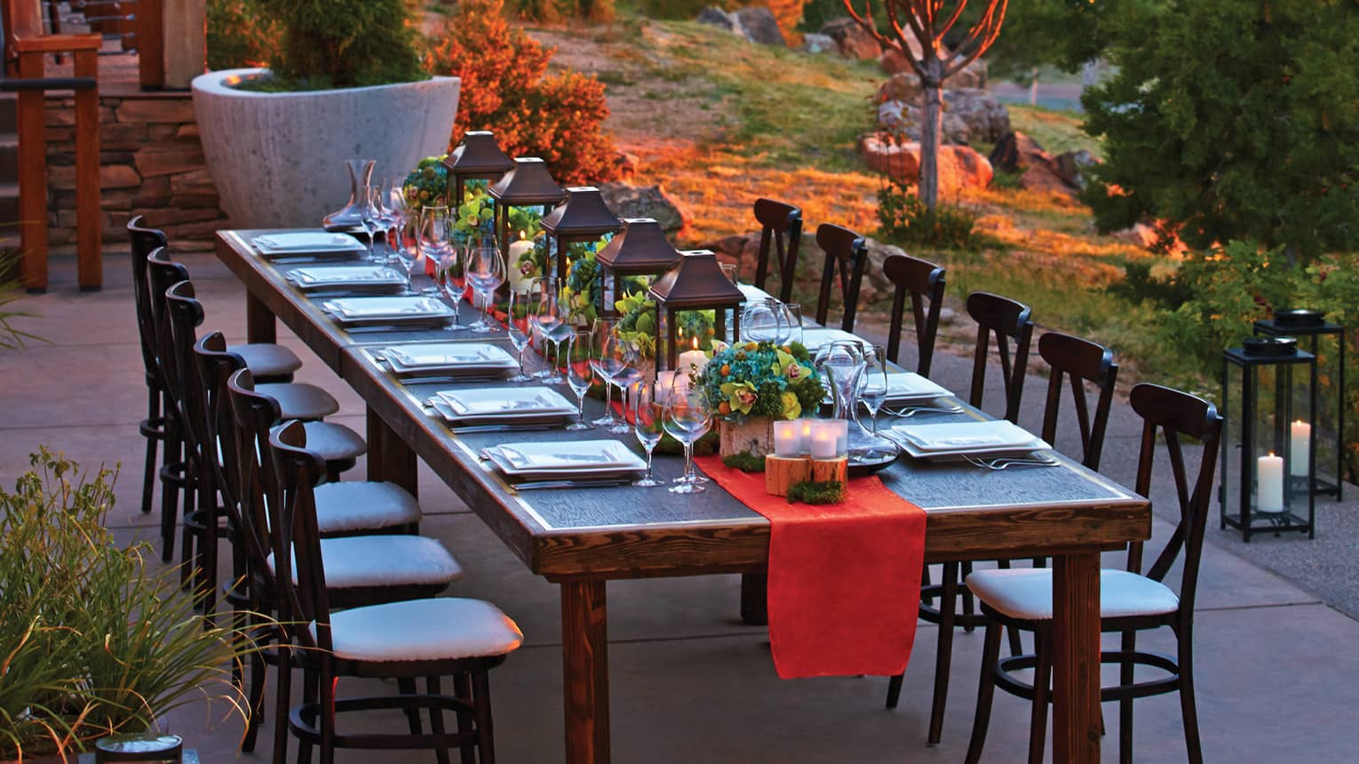 Long wooden table set with red table runner, lanterns, flowers, wine glasses on outdoor terrace at dusk