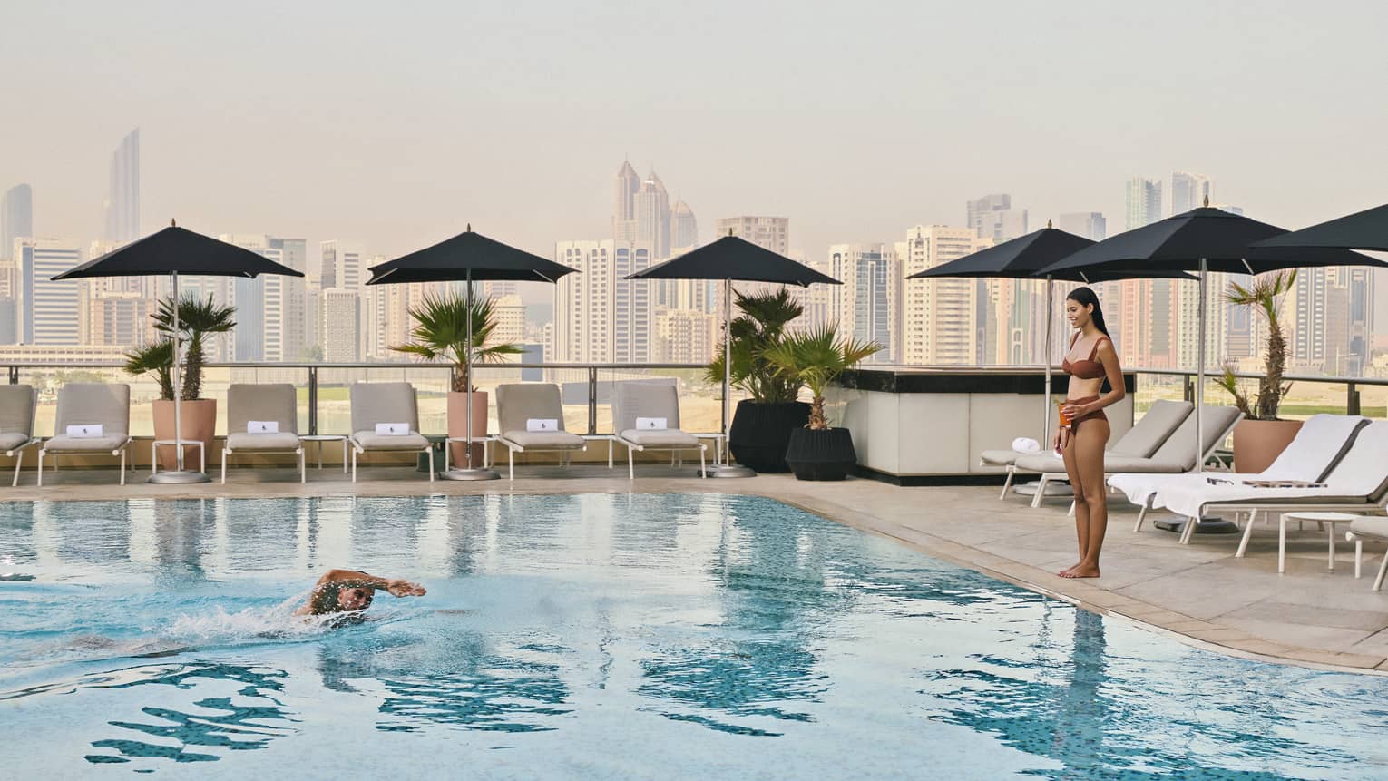 One guest stands by a rooftop pool while another swims in the water