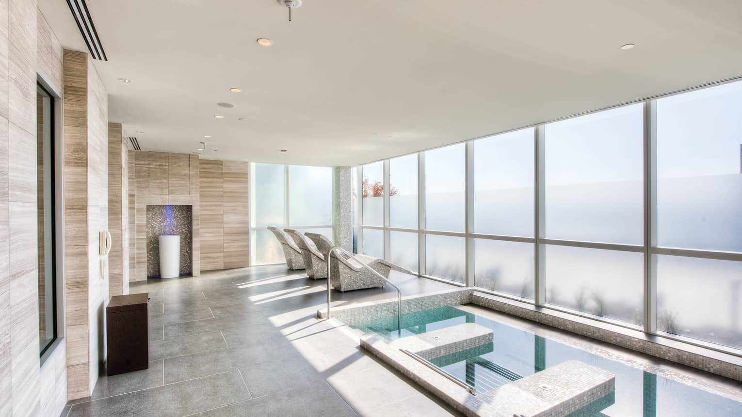 Spa plunge pool on grey tile deck, three stone chairs facing wall of windows 