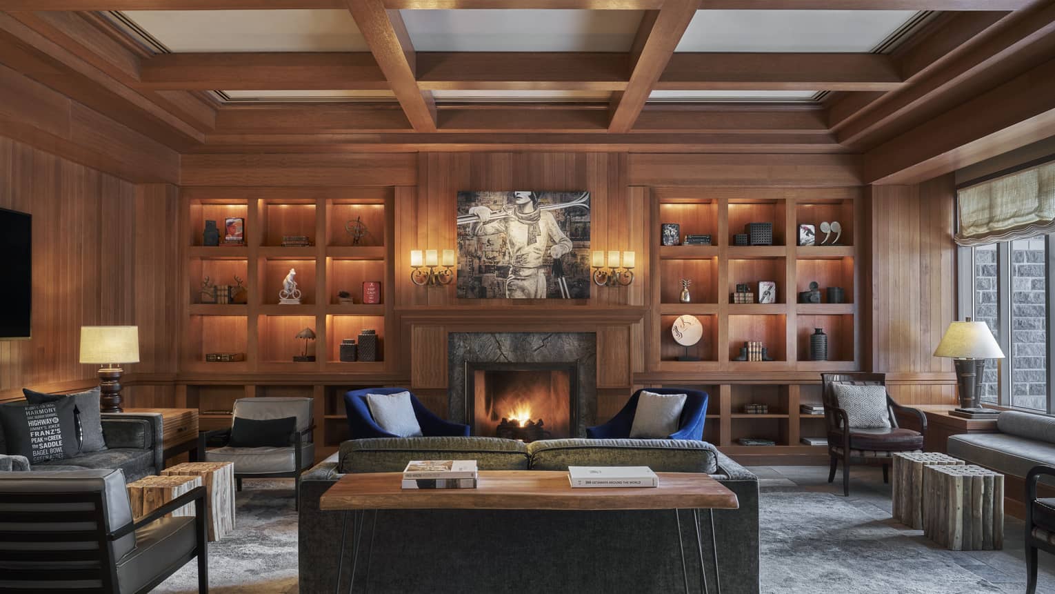 An oak library with several illuminated shelves, and a roaring fire in the stone fireplace.