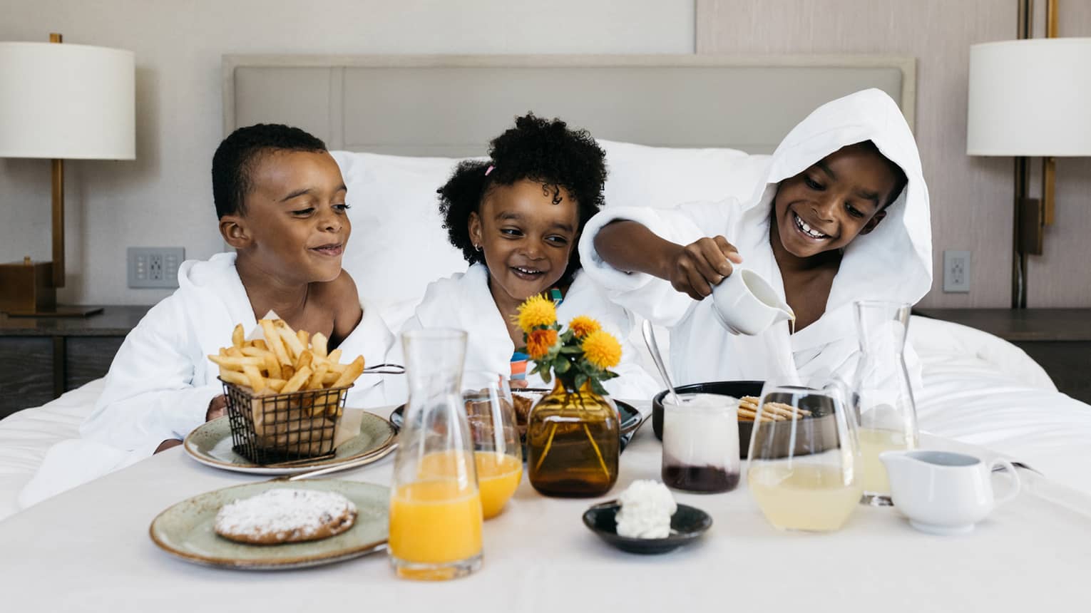 Three children in white robes enjoying breakfast in bed with fries, juice, pastries, and a flower vase at the five-star luxury hotel