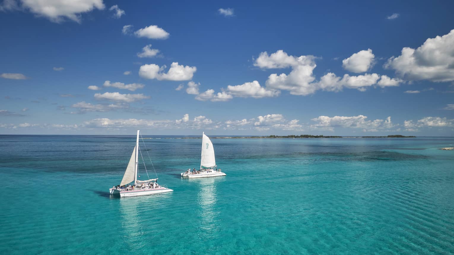 Guests aboard two catamaran sailboats glide through bright turqoise water, distant island beyond, under a cloud-dappled sky.