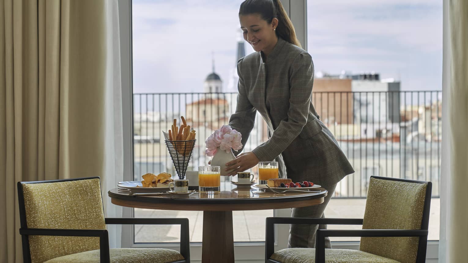 Woman places flowers on table set for breakfast for two, window with city view