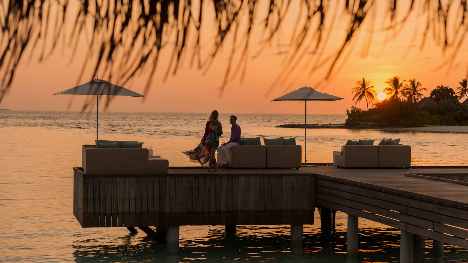 Private island dining at sunset. Woman with long dress blowing in the wind, man sits on the edge of white sofa