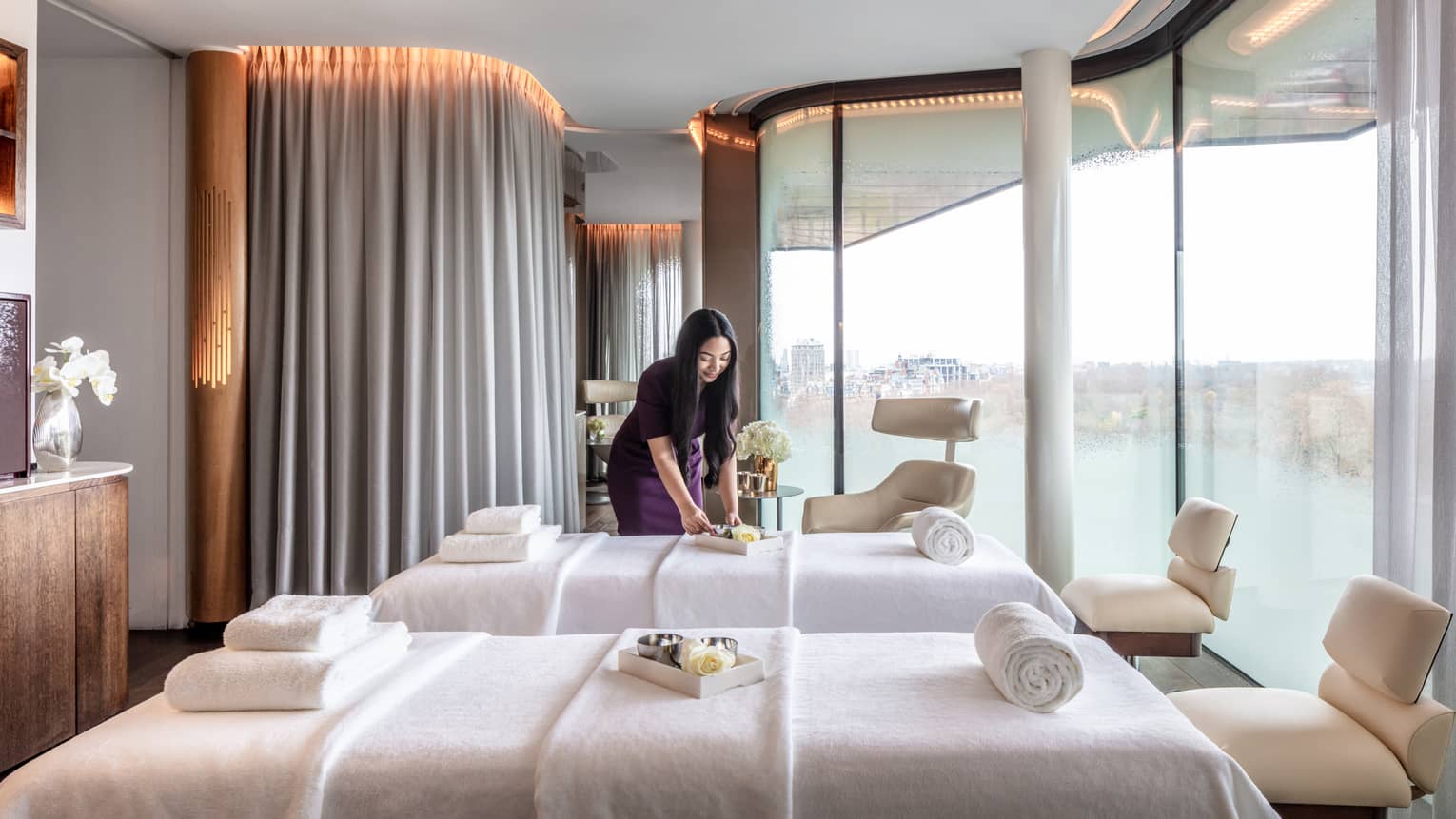 Spa couples treatment room with staff member, two white beds and chairs at ends, tall windows with city views