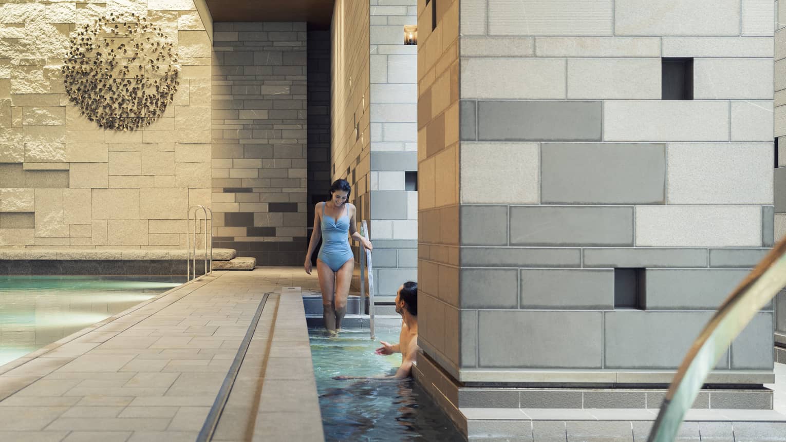 A guest enters a pool via the stairs to meet another guest who is half-hidden by a large grey-tiled column in the foreground.