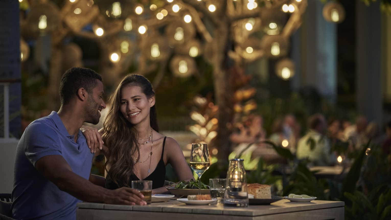 A couple sits together at a dining table beneath a tree lit with twinkling string lights