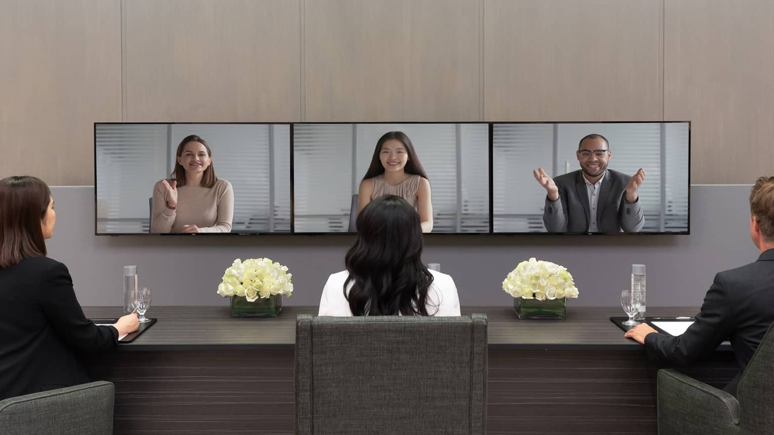 Several people in business clothes sit in front of a series of screens in a virtual meeting.