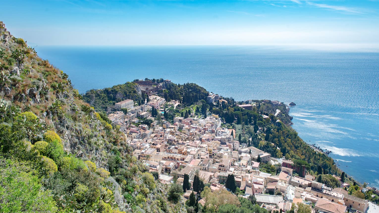 Aerial view of a coastal town with clustered buildings, lush greenery and the blue expanse of the ocean in the background.