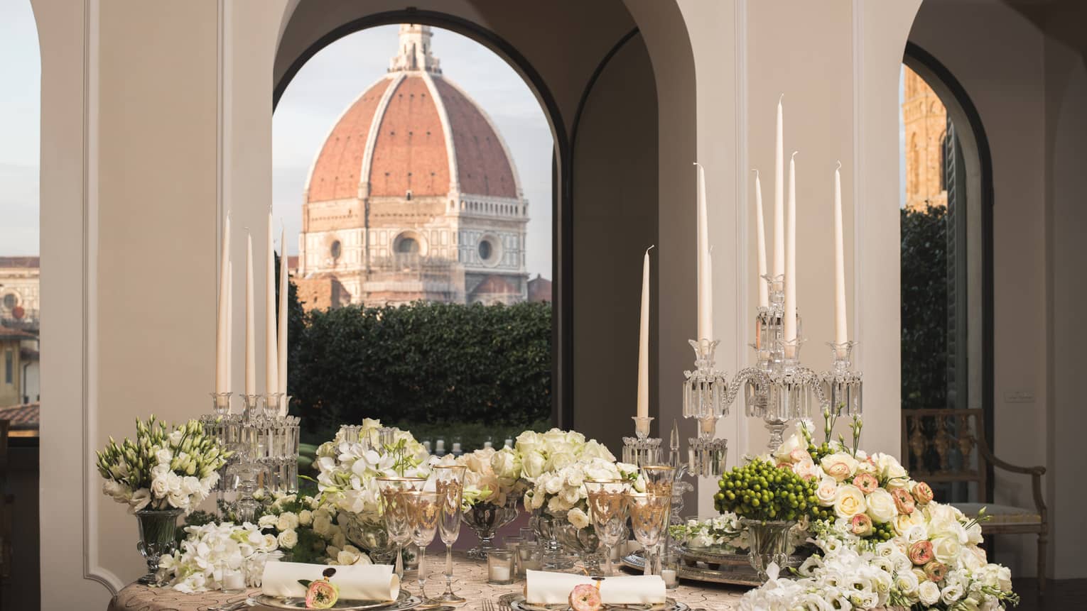 Elegant dining table set with candles, flowers, and glassware, with a large dome visible through arched windows in the background.
